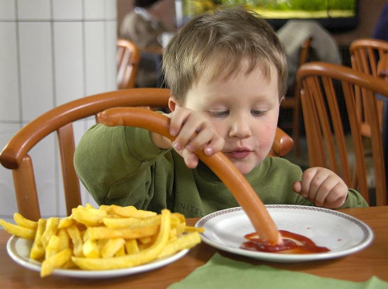 Kind isst in einem Schnellrestaurant Pommes frites und Wiener Wuerstchen mit Ketchup