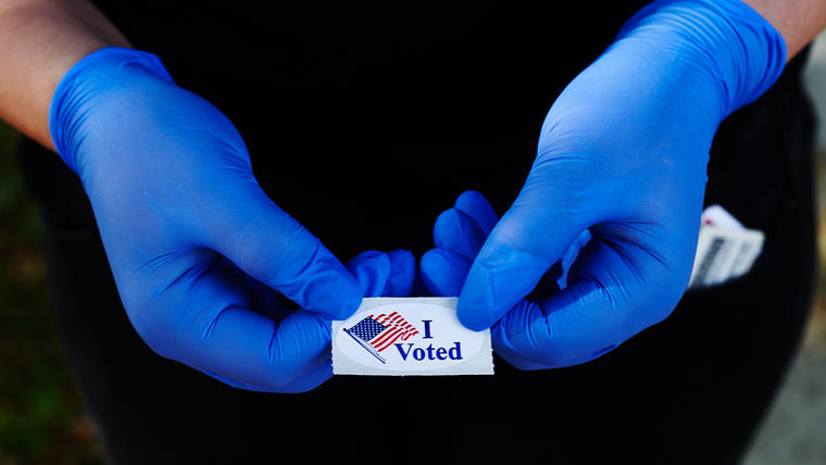 Un votante sostiene una calcomanía de "Yo voté" usando guantes de látex tras votar en las primarias de Florida, 17 de marzo en Oldsmar, Florida. Los voluntarios encuestados dicen que la participación en persona ha disminuido en la mayoría de los lugares d