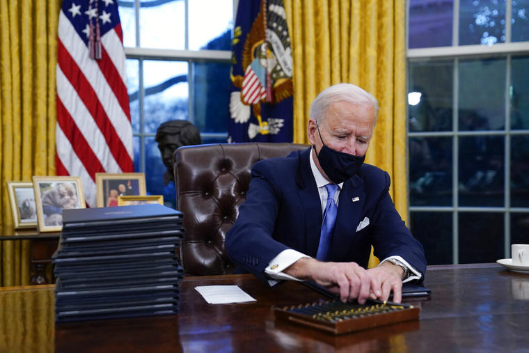 El presidente Joe Biden alcanza un bolígrafo para firmar su primera orden ejecutiva en la oficina Oval.