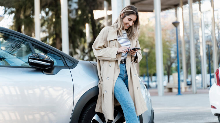 Mujer que viste con gabardina, usa su teléfono, mientras está recargada en un automóvil. 