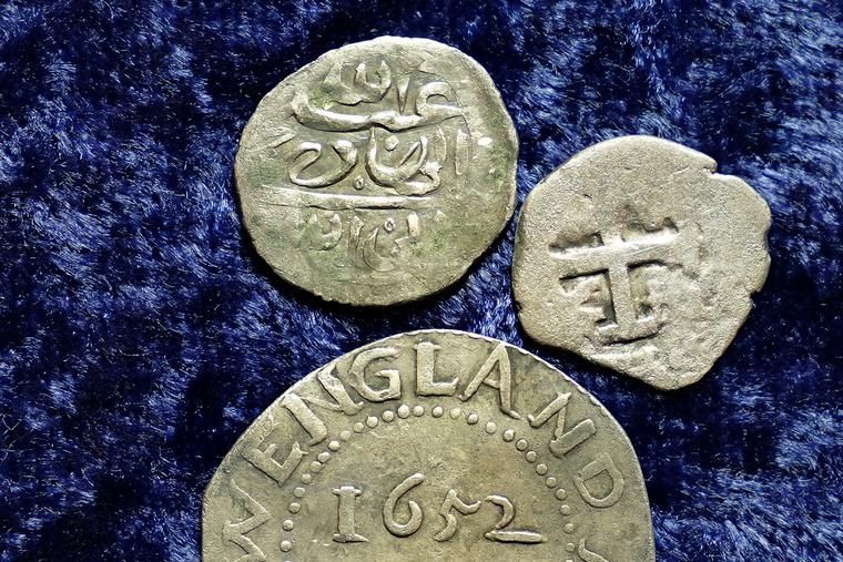 Una moneda árabe de plata del siglo XVII se aprecia en la parte superior junto a otra moneda acuñada en 1652 por la Colonia de la bahía de Massachusetts, abajo, y una moneda española de medio real de 1727, a la derecha.