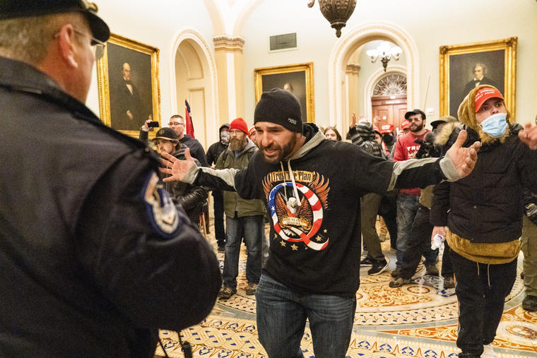 Un policía del Capitolio frente a varios seguidores de Donald Trump, ataviados con playeras a favor del presidente saliente y alusivas a la teoría de conspiración infundada Q Anon